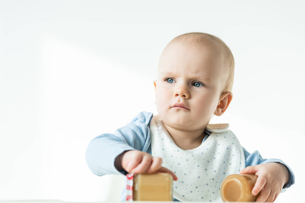 Selektiver Fokus des Babys, das Gläser mit Fruchtpüree hält und auf weißem Hintergrund wegschaut - Foto, Bild