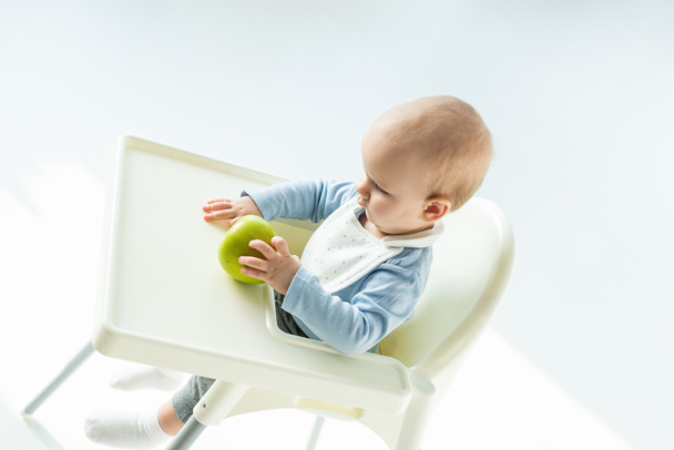 High angle view of baby boy holding green apple while sitting on feeding chair on white background - Foto, Imagen