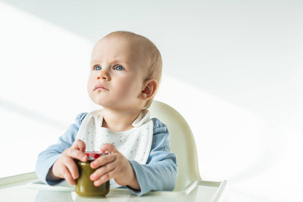 Selective focus of cute baby holding jar of vegetable baby food and looking away on feeding chair on white background - 写真・画像
