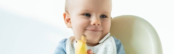 Enfoque selectivo de niño sonriente sosteniendo plátano en silla de alimentación sobre fondo blanco, tiro panorámico
 - Foto, Imagen