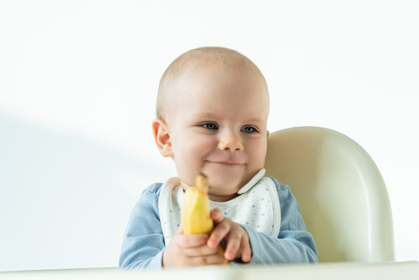 Selective focus of smiling baby holding banana while sitting on feeding chair on white background - Photo, Image