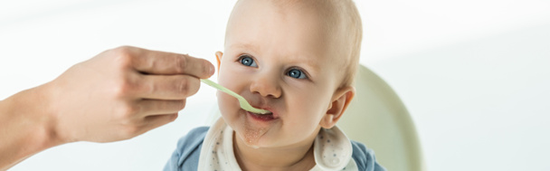 Panoramisch schot van moeder met lepel voeden schattig baby jongen op voederstoel op witte achtergrond - Foto, afbeelding