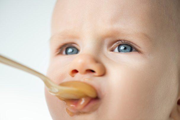 Close up view of adorable baby boy looking away during feeding with fruit puree isolated on grey  - Photo, Image