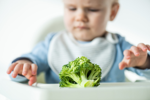 Selective focus of piece of broccoli on table near baby on feeding chair isolated on grey - Photo, Image