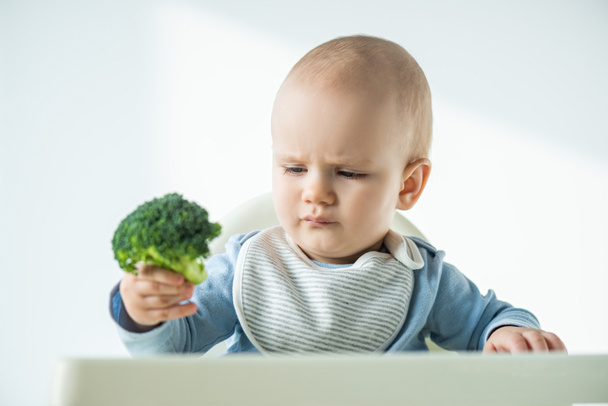 Enfoque selectivo de bebé sosteniendo pieza de brócoli mientras está sentado en la silla de alimentación sobre fondo blanco
 - Foto, imagen