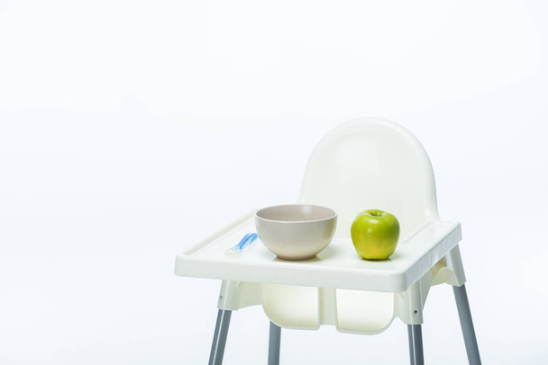 Bowl with spoon and ripe apple on table of feeding chair isolated on white - 写真・画像
