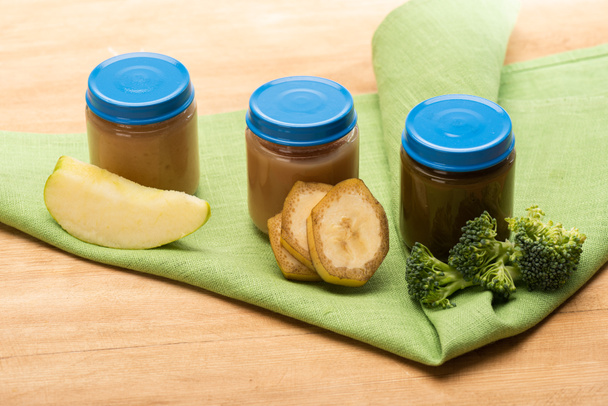 Frascos de comida para bebés con trozos de manzana, brócoli y rodajas de plátano en la servilleta en la superficie de madera
 - Foto, imagen