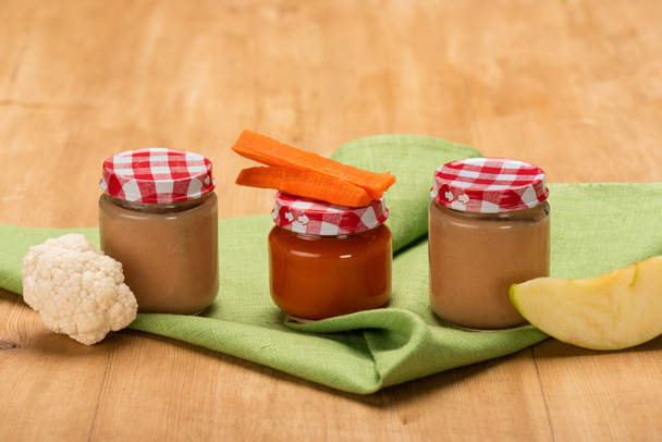 Pedazos de manzana, coliflor y zanahoria cerca de frascos de comida para bebés en la servilleta en la superficie de madera
 - Foto, Imagen