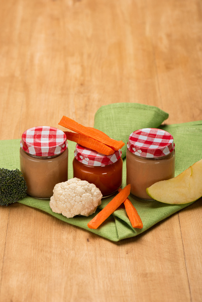 Jars of baby food with cauliflower, broccoli and carrot on napkin on wooden surface - Fotoğraf, Görsel