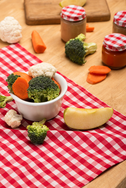 Organic vegetables and apple on napkin near jars of baby food on wooden background - Fotoğraf, Görsel