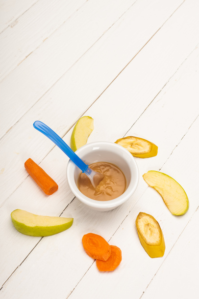 High angle view of baby food in bowl with spoon and sliced fruits and carrot on white wooden background - Fotoğraf, Görsel