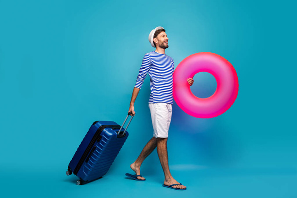 Full body profile photo of handsome guy traveler rolling case hold big pink lifebuoy want to see ocean wear striped sailor shirt cap shorts flip flops isolated blue color background - Photo, image
