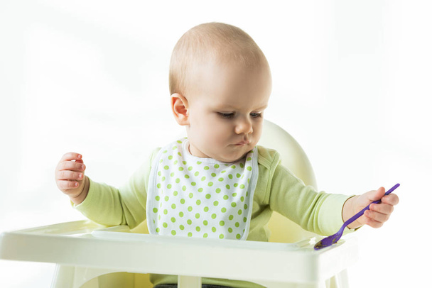 Cute baby holding spoon while sitting on feeding chair on white background - 写真・画像