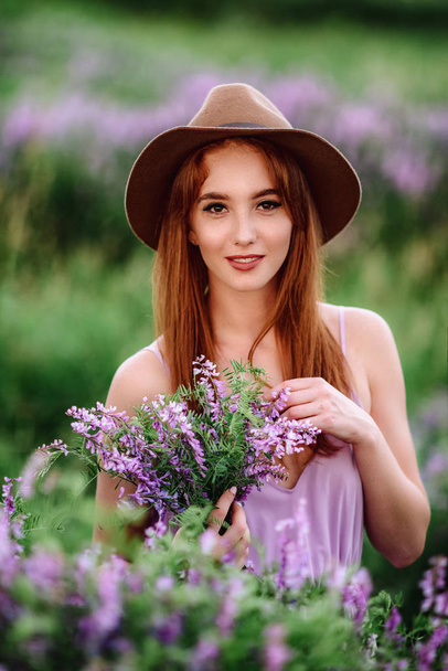 Das rothaarige Mädchen mit Hut liegt im Gras mit lila Blüten. junge Frau lächelt in der Natur. Dame geht auf Lavendelfeld. - Foto, Bild