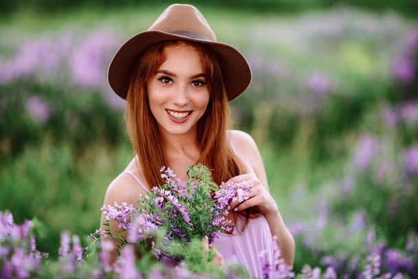 Red-haired girl in a hat lies in the grass with purple flowers. Young woman smile in nature. lady walks on a lavender field. - Photo, Image