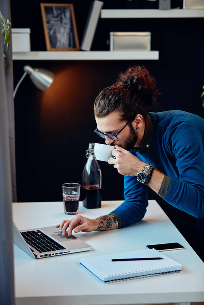 Mladý atraktivní vousatý hipster s kudrnatými vlasy opírající se o stůl, pití kávy a surfování na internetu, zatímco stojí v domácí kanceláři. - Fotografie, Obrázek