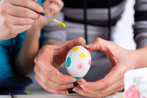 Молодая мать и ее ребенок руками рисуют пасхальные яйца. Мать помогает сыну нарисовать яйцо кистью дома. Счастливая семья готовится к Пасхе
 - Фото, изображение