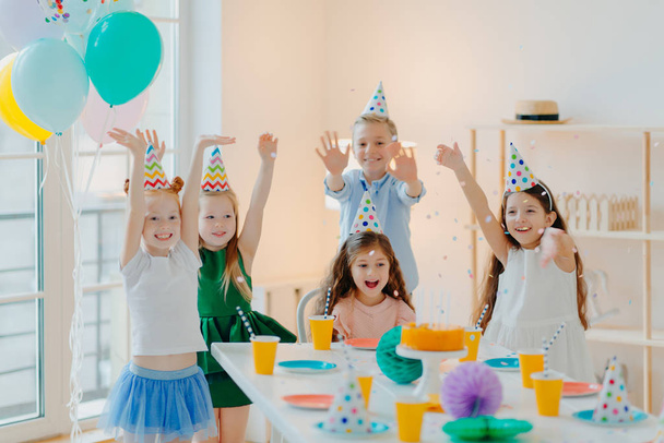 Grupa szczęśliwych dzieci świętować urodziny razem, grać z konfetti, nosić kapelusze party, pozować w pobliżu świątecznego stołu w urządzonym pokoju z balonami, mają overjoyed wyrażenia, cieszyć się życiem - Zdjęcie, obraz