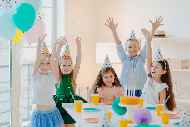 Feliz fiesta de cumpleaños. Joyful small children have fun together, raise arms and play with confetti, pose at festive table, wear party hats, being on carnival, enjoy holidays. - Foto, imagen