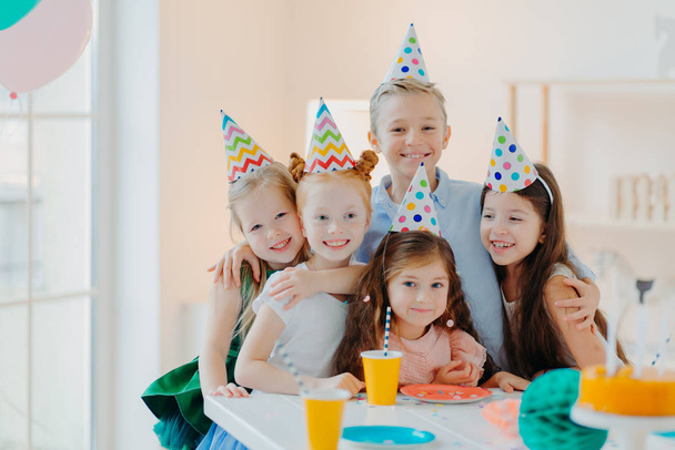 Öt barátságos kisgyerek visel ünnepi kúp sapkát, ölelés és fotózás együtt, játék és születésnapi ünneplés, boldog kifejezések, pózolni ünnepi asztalnál - Fotó, kép