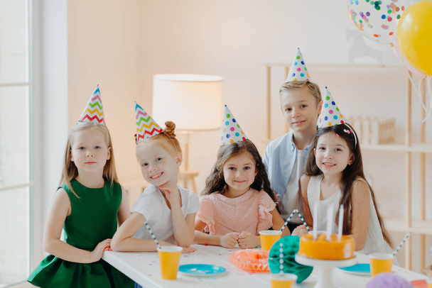 Crianças, celebração e conceito de aniversário. Crianças positivas se divertir juntos na festa, usar chapéus cone, comer bolo delicioso, jogar jogos interessantes, estar em bom humor - Foto, Imagem