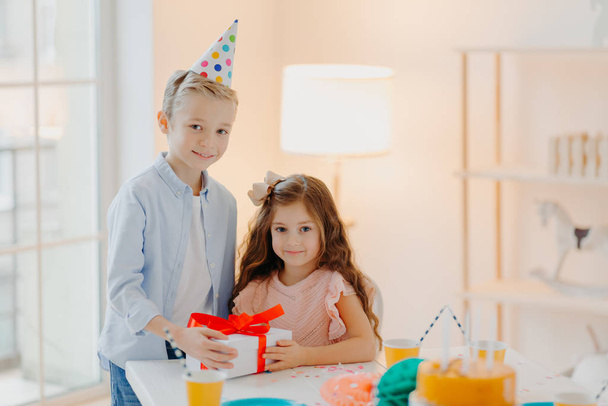 Handsome small boy gives present box to girl, celebrate birthday together, wear festive clothes and party cone hats, pose at table with cake in white spacious room. Holiday concept - Photo, Image