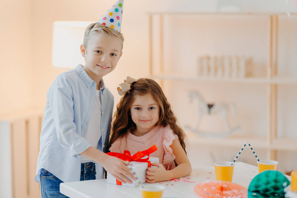 Happy small girl and boy hold gift box with red ribbon, prepare surprise on birthday, pose at white table, wear cone party hats, have positive expressions. Childhood concept - Photo, Image