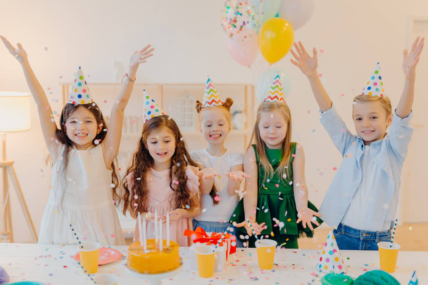 Tiro horizontal de crianças felizes positivo pegar confete, celebrar o aniversário juntos, levantar os braços, ter bom humor, jogar juntos, ficar perto da mesa festiva com caixa presente, bolo, chapéus de festa - Foto, Imagem