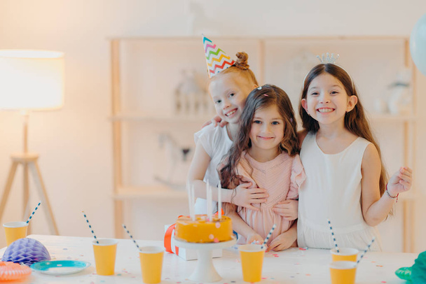 Horizontal shot of three happy friends embrace and have funny expressions, pose near festive table with cake indoor. Three girls celebrate birthday together, have fun, come to party - Photo, Image