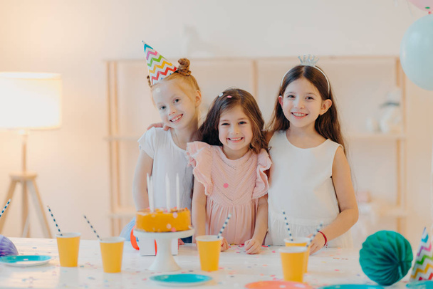 Внутренний снимок счастливых трех девочек, которые обнимаются и веселятся, радостно улыбаются, стоят возле праздничного стола с тортом, чашками, счастливо проводят детство, находясь на вечеринке вместе. Концепция детства и праздника - Фото, изображение