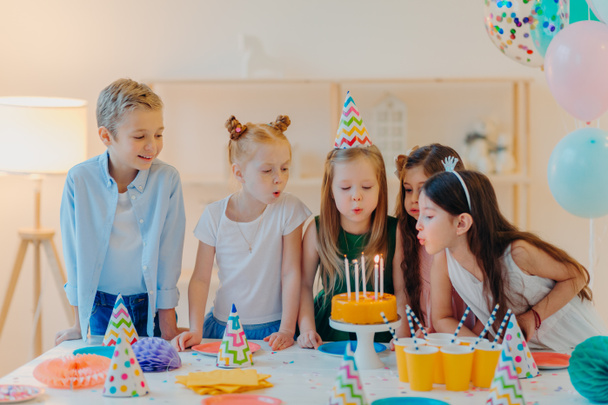 Small kids celebrate birthday party, blow candles on cake, gather at festive table, have good mood, enoy spending time together, make wish, wear party hats, pose indoor with inflated balloons - Photo, Image