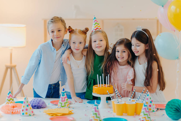 Friendly children embrace while pose near festive table, blow candles on cake, have party mood, celebrate birthday or special occasion, have joyful expressions. Childhood, fun and entertainment - Photo, Image