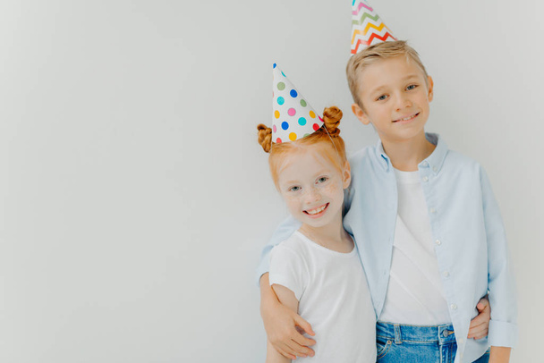 Happy small jinger freckled girl and little boy embrace and smile gladfully, wear party hats, enjoy nice time together, isolated over white background, copy space. Concepto infantil y festivo de eventos - Foto, imagen