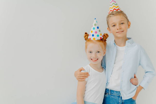 Isolated shot of happy broher and sister cuddle each other, have positive expressions, wear party hats, going to celebrate birthday, stand against white background, copy space aside. Niños alegres - Foto, imagen