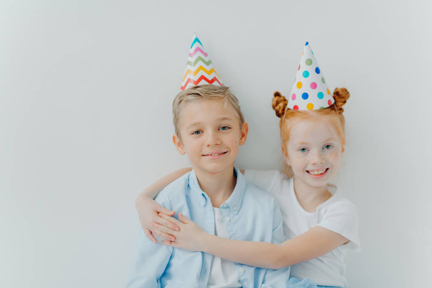 Lovey red haird girl embraces with love her older brother, congratulates with birthday, wear cone party hats, have good mood on party, isolated over white background, have friendly relationship - Photo, Image
