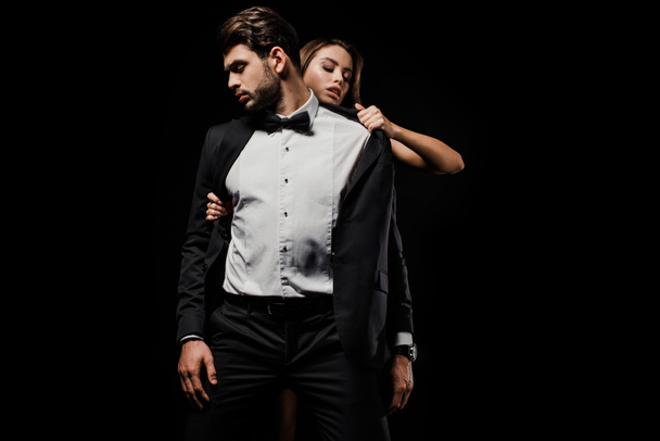 passionate woman taking off jacket of handsome man in suit isolated on black  - Fotoğraf, Görsel