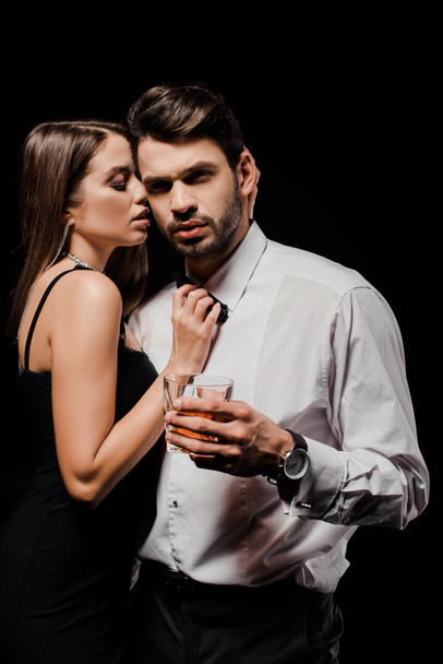 σέξι γυναίκα που αγγίζει παπιγιόν του όμορφου άνδρα με ένα ποτήρι ουίσκι απομονωμένο σε μαύρο  - Φωτογραφία, εικόνα