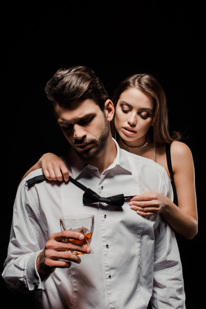 attractive woman holding bow tie near man with glass of whiskey isolated on black  - Фото, изображение
