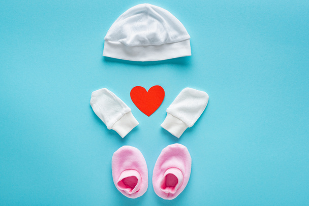 Vue du dessus des mitaines, chapeau et chaussons bébé avec coeur en papier sur fond bleu, concept de fête des mères
  - Photo, image
