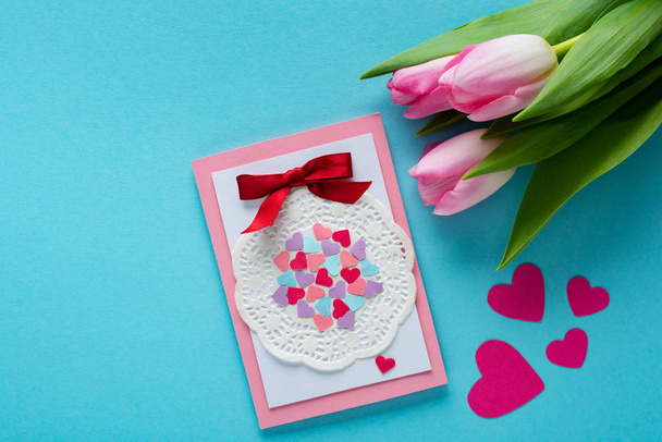 Top view of greeting card with tulips and paper hearts on blue surface - 写真・画像