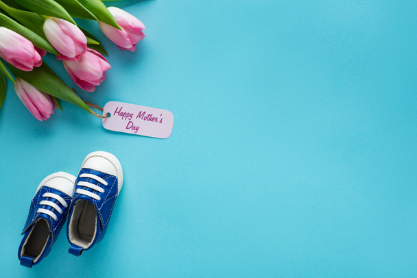 Vue du dessus des tulipes avec le lettrage heureux de la fête des mères sur l'étiquette en papier près des chaussons de bébé sur fond bleu
 - Photo, image