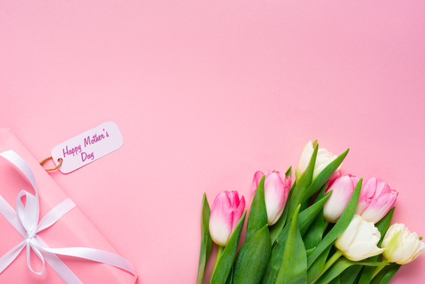 Top view of gift with happy mothers day lettering on paper label near tulips on pink background - Foto, Bild