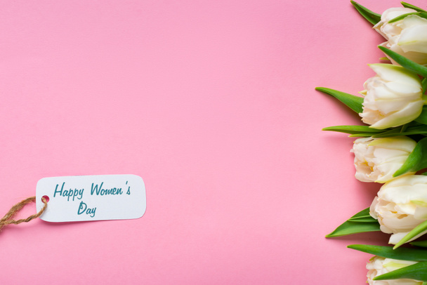 Vista dall'alto di etichetta di carta con felice giorno delle donne lettering vicino tulipani bianchi su sfondo rosa
 - Foto, immagini