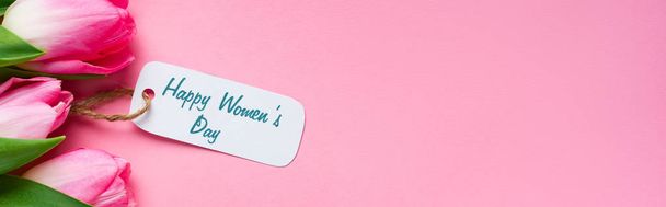 Top view of happy womens day lettering on paper label with tulips on pink surface, panoramic shot  - Foto, imagen