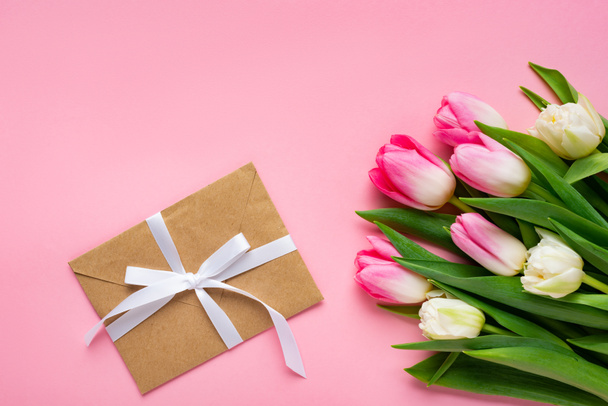 Top view of envelope with bow and bouquet of tulips on pink surface - Photo, Image