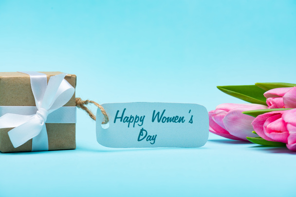 Caixa de presente com letras felizes do dia das mulheres no rótulo de papel e tulipas no fundo azul
 - Foto, Imagem