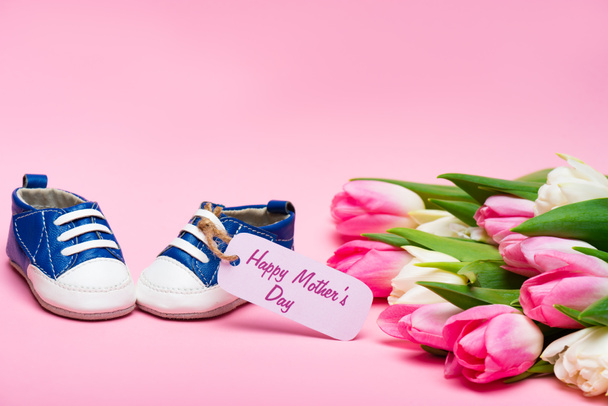 Botines de bebé con letras felices del día de las madres en la etiqueta de papel cerca del ramo de tulipanes en la superficie rosa
 - Foto, imagen