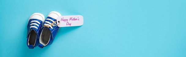 Vue du dessus des chaussons pour bébés avec lettrage heureux de la fête des mères sur l'étiquette en papier sur la surface bleue, vue panoramique
 - Photo, image
