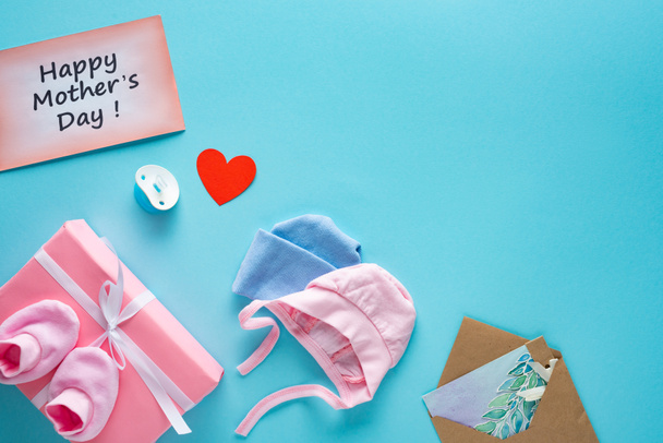 Vista superior de la caja de regalo, ropa de bebé y tarjetas de felicitación con letras felices del día de las madres en la superficie azul
 - Foto, Imagen