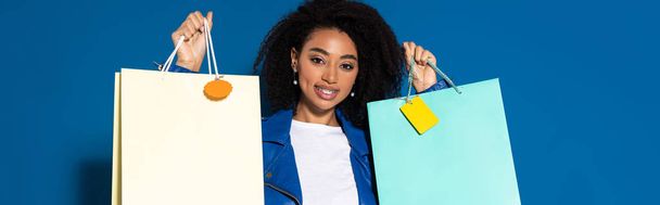 sorrindo bela mulher afro-americana segurando sacos de compras no fundo azul, tiro panorâmico
 - Foto, Imagem